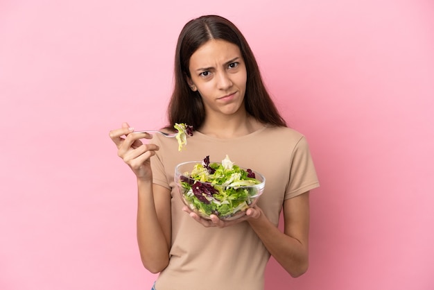 Jovem francesa isolada em um fundo rosa segurando uma tigela de salada com uma expressão triste