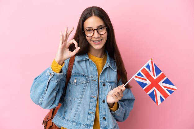 Jovem francesa segurando uma bandeira do reino unido isolada em um fundo rosa, mostrando o sinal de ok com os dedos