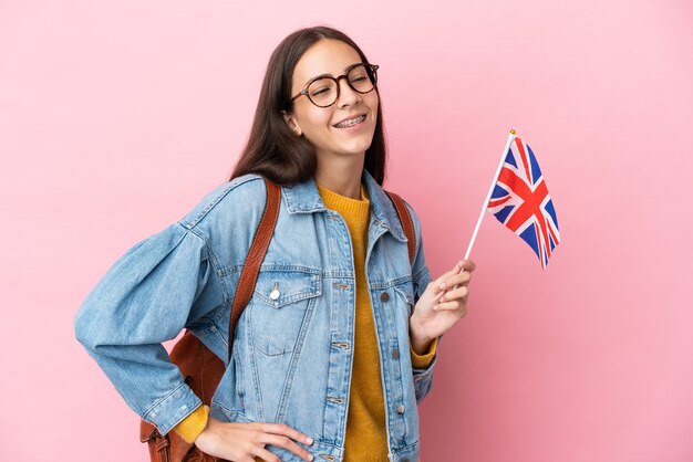 Jovem francesa segurando uma bandeira do Reino Unido isolada em um fundo rosa, posando com os braços na cintura e sorrindo
