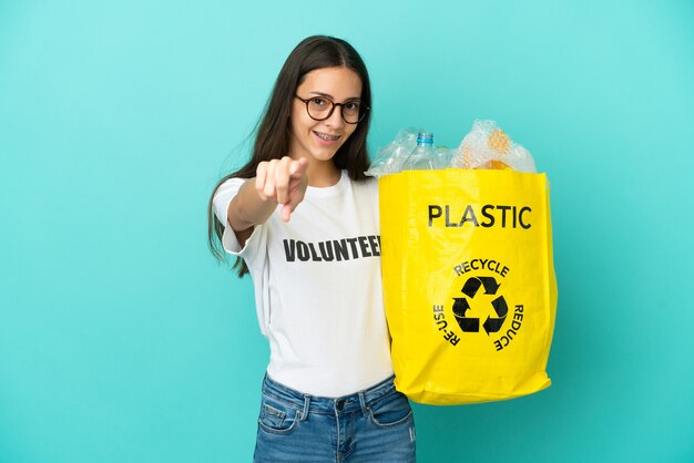 Foto jovem francesa segurando uma sacola cheia de garrafas plásticas para reciclar apontando para a frente com uma expressão feliz