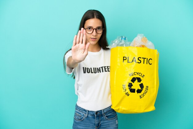 Jovem francesa segurando uma sacola cheia de garrafas plásticas para reciclar, fazendo um gesto de pare