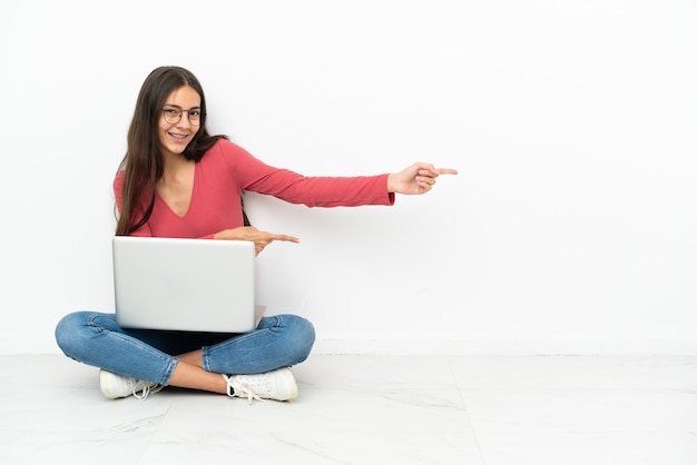 Jovem francesa sentada no chão com seu laptop apontando o dedo para o lado e apresentando um produto