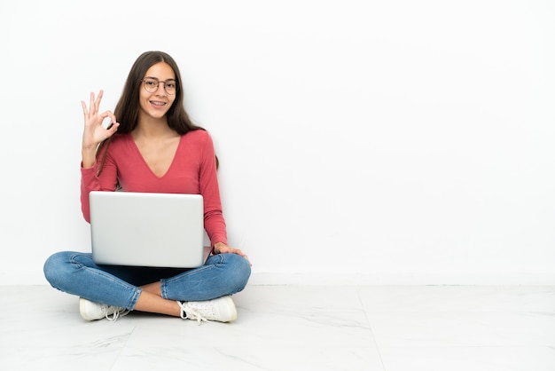 Jovem francesa sentada no chão com seu laptop, mostrando um sinal de ok com os dedos