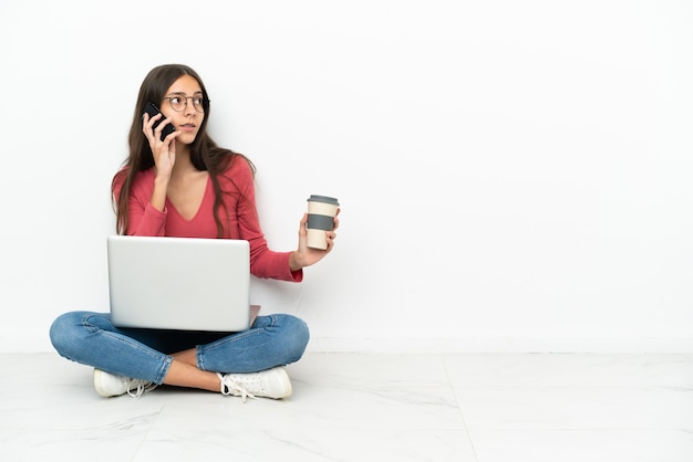 Jovem francesa sentada no chão com seu laptop segurando um café para levar e um celular