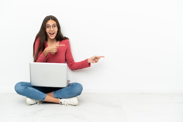 Jovem francesa sentada no chão com seu laptop surpresa e apontando para o lado