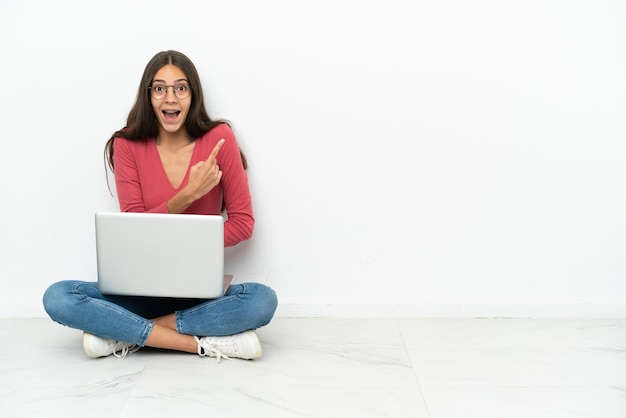 Jovem francesa sentada no chão com seu laptop surpresa e apontando para o lado