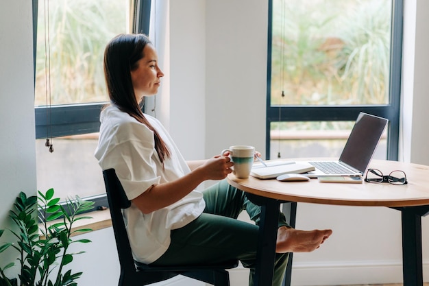 Jovem freelancer a beber café e a trabalhar no portátil em casa.