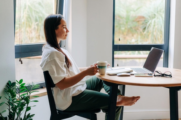 Foto jovem freelancer a beber café e a trabalhar no portátil em casa.