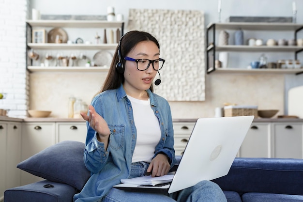 Jovem freelancer asiática trabalhando remotamente em casa usando fone de ouvido e laptop para videochamada