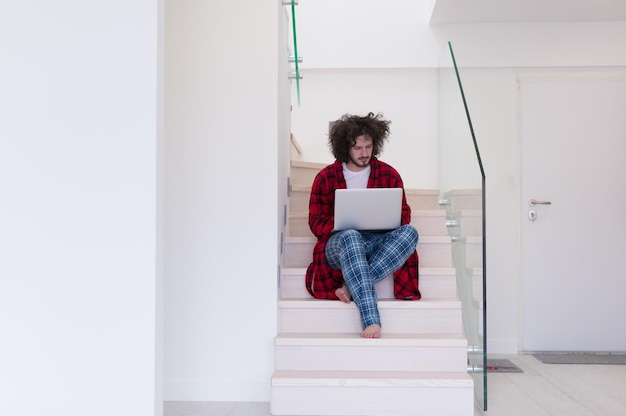 jovem freelancer em roupão trabalhando em casa usando o laptop enquanto está sentado na escada