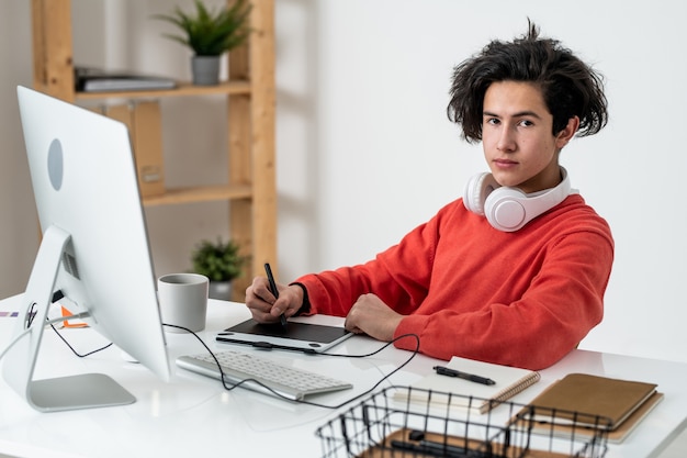 Jovem freelancer masculino casual com fones de ouvido olhando para você enquanto retoca fotos em uma mesa digitalizadora em casa