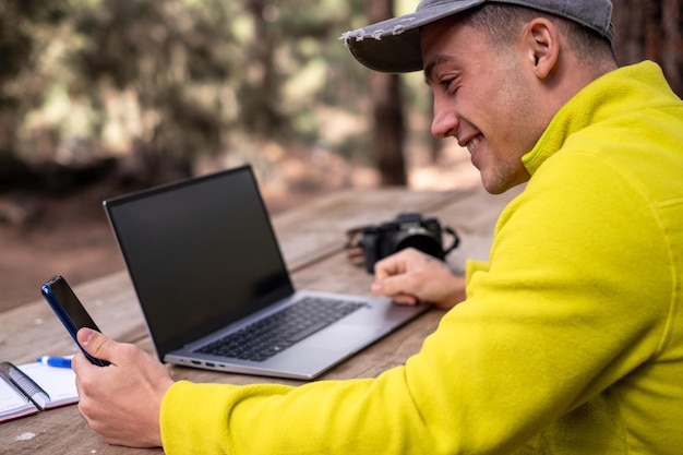 Jovem freelancer masculino caucasiano usando telefone celular enquanto trabalhava no computador portátil Viajante remoto Hipster menino trabalhando distante enquanto aprecia a paisagem natural durante as férias