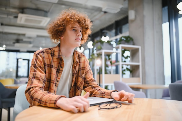 Jovem freelancer masculino feliz em roupas casuais sentado no café com laptop e usando telefone celular