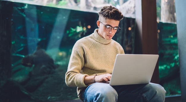 Jovem freelancer masculino inteligente em óculos e roupas casuais digitando no laptop enquanto trabalha em ambientes fechados