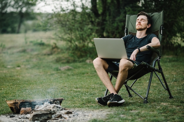 Foto jovem freelancer relaxante na floresta. homem trabalhando no laptop na natureza. trabalho remoto, atividade ao ar livre no verão. viagens, caminhadas, tecnologia, turismo, conceito de pessoas - homem sentado na cadeira ao ar livre.