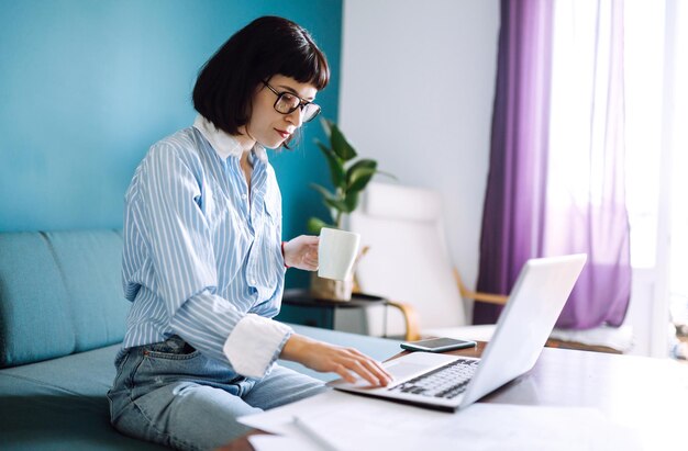 Jovem freelancer senta-se no sofá moderno e trabalha no laptop. Educação on-line. Trabalhando em casa.