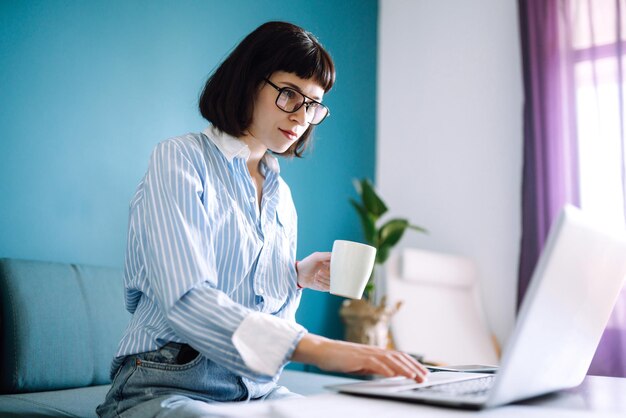 Jovem freelancer senta-se no sofá moderno e trabalha no laptop. Educação on-line. Trabalhando em casa.