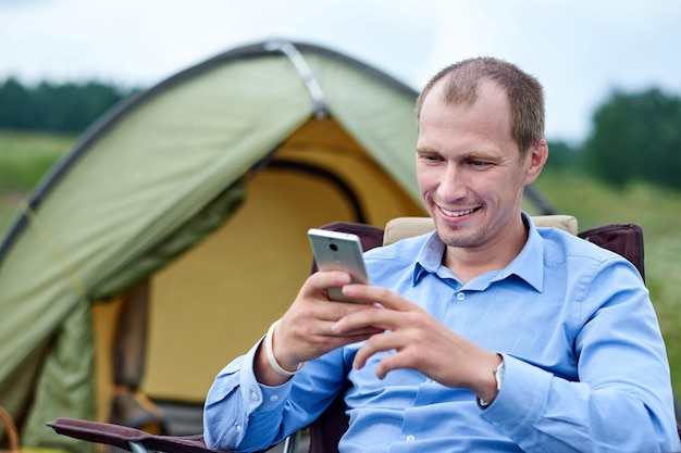 Jovem freelancer sentado na cadeira e usando telefone inteligente Relaxando na frente da barraca no acampamento na floresta ou prado Trabalho remoto e atividade ao ar livre no verão