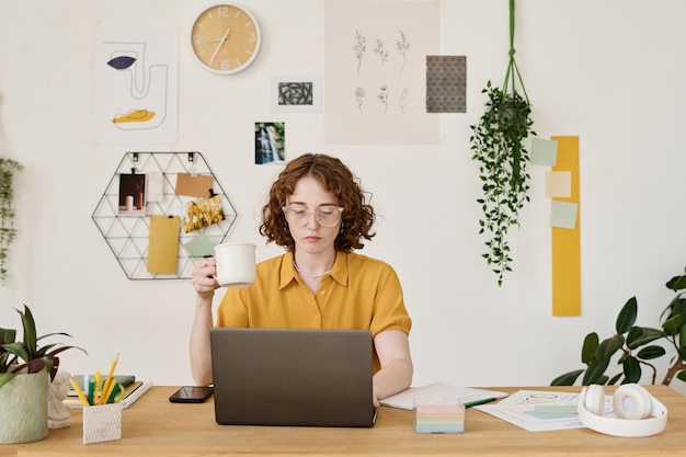 Jovem freelancer sério com uma xícara de chá sentado na frente do laptop pelo local de trabalho