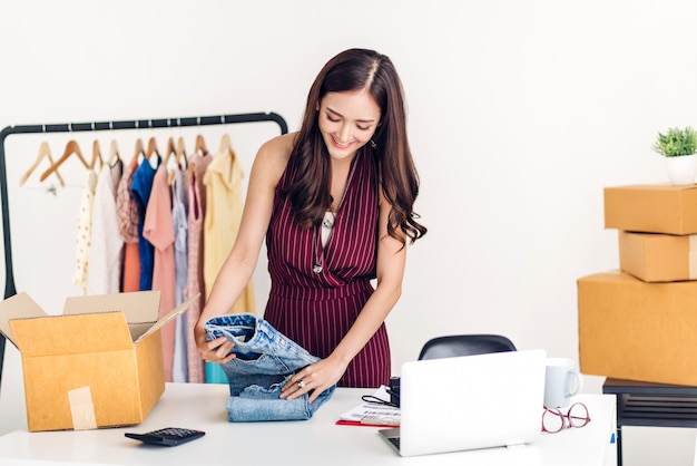 Jovem freelancer trabalhando sme negócios compras on-line e embalagem de roupas com caixa de papelão em casa - conceito de transporte e entrega on-line de negócios