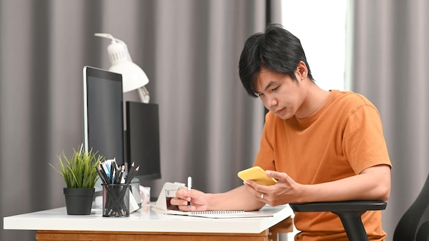 Jovem freelancer usando telefone inteligente e anotando no notebook
