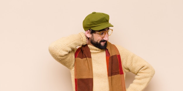 Jovem frio barbudo sorrindo alegre e casualmente, levando a mão na cabeça com um olhar positivo, feliz e confiante