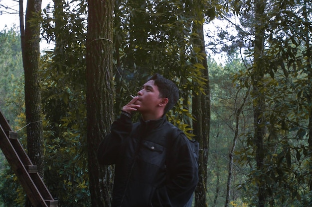 Foto jovem fumando enquanto está de pé perto de árvores na floresta