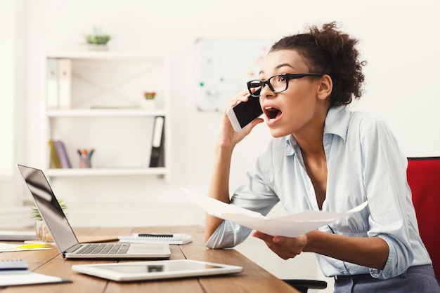 Jovem furiosa mulher afro-americana, falando no telefone no espaço da cópia do escritório moderno. Consultoria de negócios.