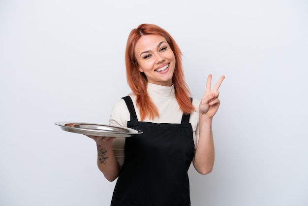 Foto jovem garçom russo com bandeja isolada no fundo branco sorrindo e mostrando sinal de vitória