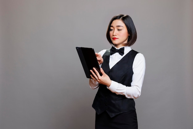 Jovem garçonete asiática de uniforme verificando o menu do café no tablet digital. Equipe de serviço de catering gerenciando pedidos on-line e tocando na tela sensível ao toque enquanto posava no estúdio em fundo cinza