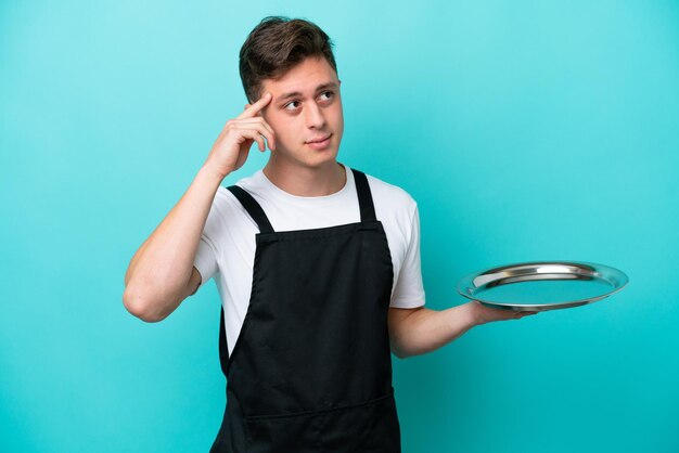 Jovem garçonete com bandeja isolada em fundo azul com dúvidas e pensando