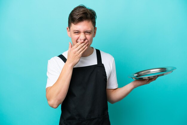 Jovem garçonete com bandeja isolada em fundo azul feliz e sorridente cobrindo a boca com a mão