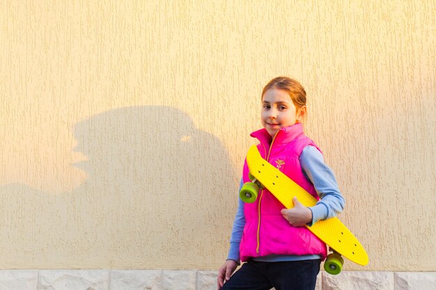 Jovem garota adorável e sorridente do lado de fora perto da parede segurando a placa de moeda amarela debaixo do braço olhando para a câmera Garota posando alegremente segurando seu novo skate atual