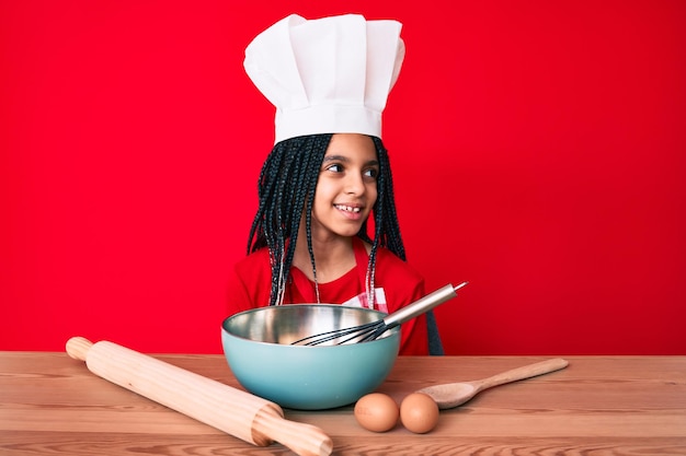 Jovem garota afro-americana com tranças usando avental de cozinheiro profissional olhando para o lado relaxar pose de perfil com rosto natural e sorriso confiante