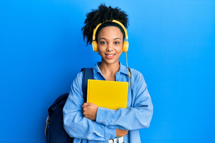 Foto jovem garota afro-americana usando mochila de estudante e fones de ouvido segurando livro sorrindo com um sorriso feliz e legal no rosto mostrando os dentes