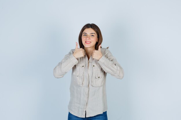 Jovem garota aparecendo dois polegares para cima em uma camisa bege, jeans e olhando alegre, vista frontal.