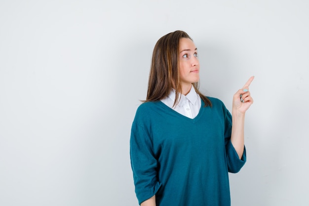 Foto jovem garota apontando para cima com o dedo em uma camisola com decote em v, camisa e olhando curiosa, vista frontal.