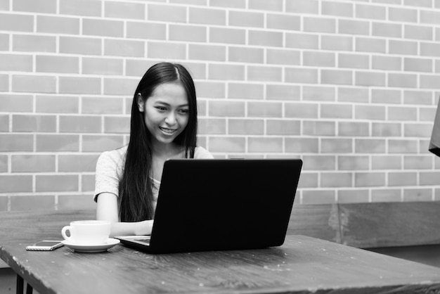 Jovem garota asiática feliz sorrindo enquanto usa o laptop com cappuccino e o celular na mesa de madeira na parede de tijolos