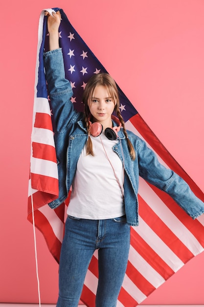 Jovem garota atraente com duas tranças na jaqueta jeans e jeans com fones de ouvido no pescoço segurando a grande bandeira americana nas mãos enquanto sonhadoramente olhando na câmera sobre fundo rosa