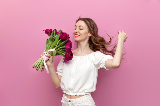 Jovem garota bonita em roupas festivas segura um buquê de flores e sorri em fundo rosa