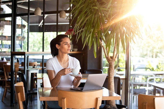 Jovem garota bonita sentada em uma mesa em um café perto da janela bebendo café cappuccino Mulher bonita trabalhando com laptop de uma cafeteria Mulher atraente sentada em um café com um laptop