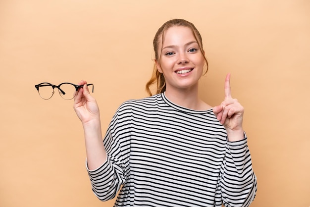 Foto jovem garota caucasiana com óculos isolados em fundo bege apontando uma ótima ideia