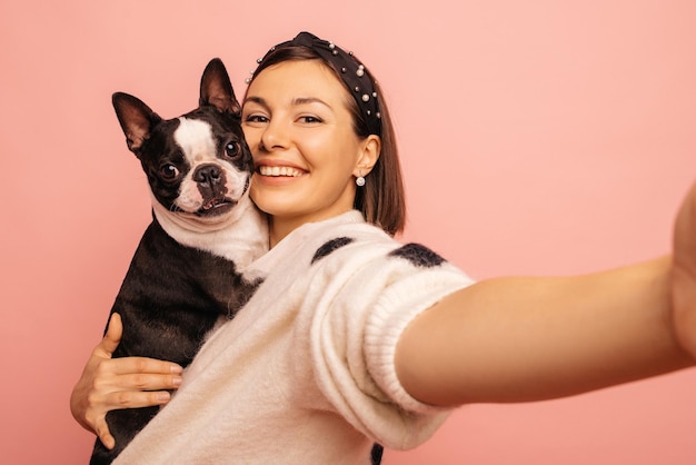 Jovem garota caucasiana com sorriso sincero tira selfie com cachorro preto e branco nos braços no fundo rosa Conceito de animais de estimação