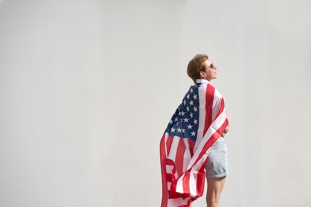 Jovem garota caucasiana, envolvida na bandeira dos EUA, olhando para longe, fundo da parede cinza