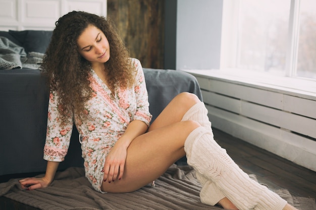 Jovem garota caucasiana sentada no chão perto da cama, vestida com um roupão de banho e meias quentes