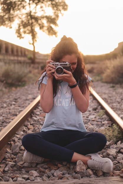 Jovem garota com cabelo comprido, sentada nos trilhos com câmera analógica, tirando fotos ao pôr do sol. Conceito de viagens.