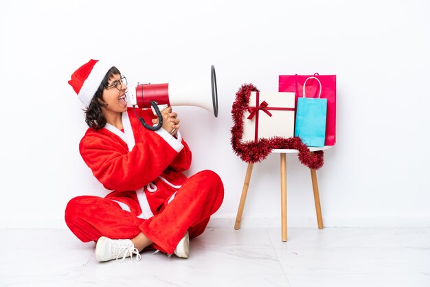 Jovem garota comemorando o natal sentada no chão, isolada em um fundo branco, gritando no megafone