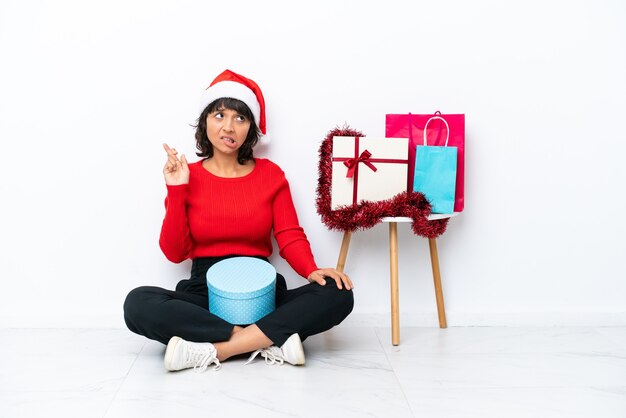 Jovem garota comemorando o natal sentada no chão, isolada no fundo branco com os dedos se cruzando e desejando o melhor