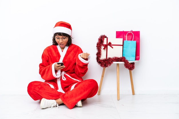 Jovem garota comemorando o natal sentada no chão, isolada no fundo branco, surpresa e enviando uma mensagem