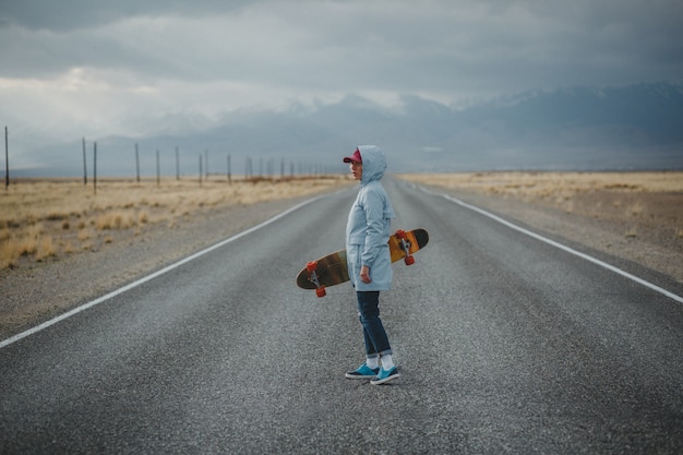 Jovem garota desportiva com longboard está de pé em uma estrada deserta